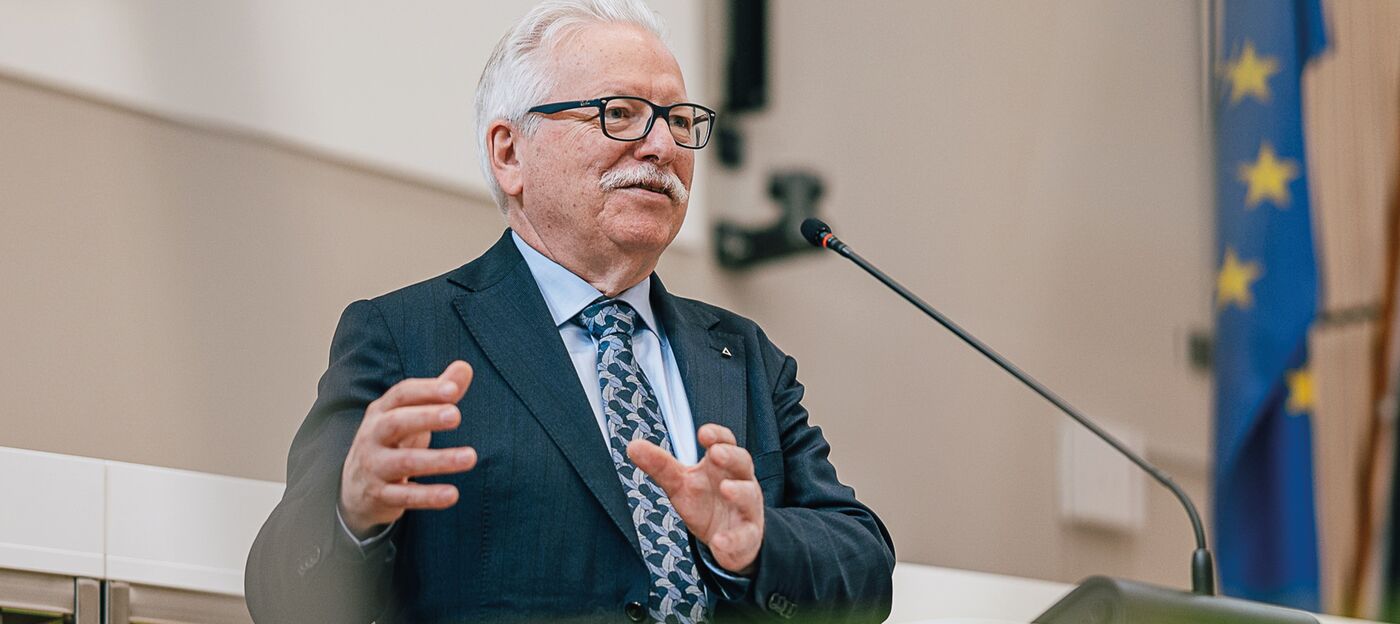 Przy mównicy stoi mężczyzna w okularach i garniturze